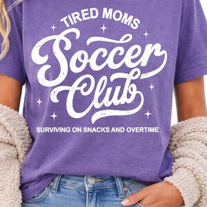 May include: A purple t-shirt with the text "TIRED MOMS Soccer Club" in white script. The phrase "SURVIVING ON SNACKS AND OVERTIME" is printed below. The shirt is worn with blue jeans and a beige cardigan.