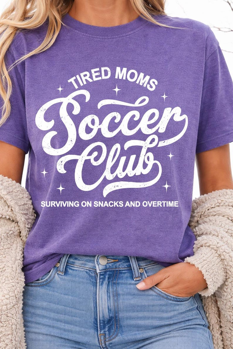 May include: A purple t-shirt with the text "TIRED MOMS Soccer Club" in white script. The phrase "SURVIVING ON SNACKS AND OVERTIME" is printed below. The shirt is worn with blue jeans and a beige cardigan.
