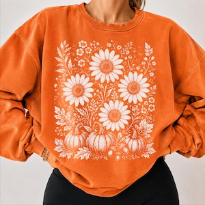 May include: An orange crewneck sweatshirt featuring a white graphic print of sunflowers, pumpkins, and foliage. The design is centered on the chest, showcasing detailed botanical illustrations of flowers and gourds in a clean, white line art style.