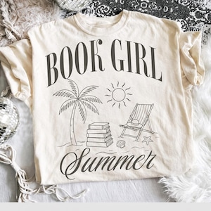 May include: Cream-colored t-shirt with a black graphic design. The design includes the words "BOOK GIRL Summer" with illustrations of a palm tree, sun, books, beach chair, hat, seashell, and starfish.