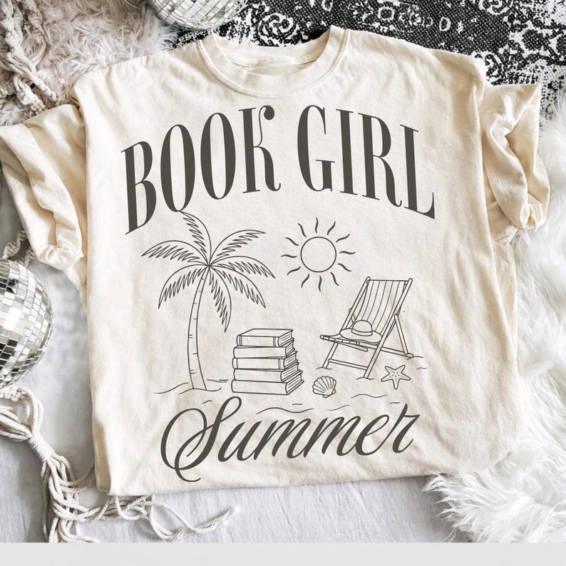 May include: Cream-colored t-shirt with a black graphic design. The design includes the words "BOOK GIRL Summer" with illustrations of a palm tree, sun, books, beach chair, hat, seashell, and starfish.