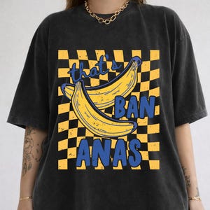 May include: Black t-shirt with a yellow and black checkered pattern. The shirt features a graphic of two yellow bananas and the text "that's BANANAS" in blue. The shirt has short sleeves and a crew neck.