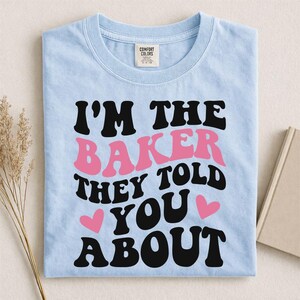 May include: Light blue t-shirt with the text "I'M THE BAKER THEY TOLD YOU ABOUT" in black and pink lettering. Two pink hearts are also on the shirt. The shirt is folded on a light surface.