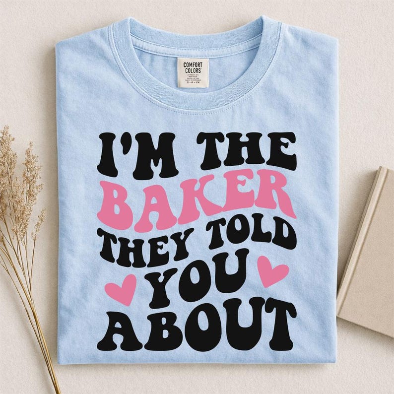 May include: Light blue t-shirt with the text "I'M THE BAKER THEY TOLD YOU ABOUT" in black and pink lettering. Two pink hearts are also on the shirt. The shirt is folded on a light surface.