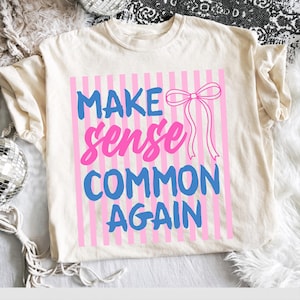 May include: Cream-colored t-shirt with the text "MAKE sense COMMON AGAIN" in blue and pink. The text is set against a pink striped background with a pink bow. The shirt is a casual wear item.