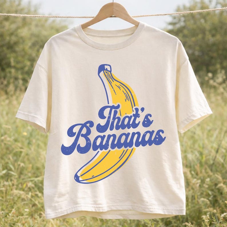 May include: Cream-colored t-shirt with a yellow banana graphic and the blue text "That's Bananas." The t-shirt is hanging on a wooden hanger against a blurred outdoor background. The shirt has short sleeves and a crew neck.