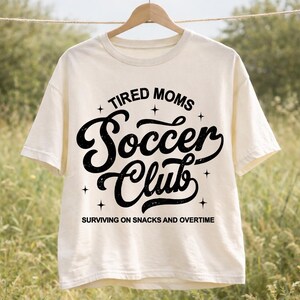 May include: Cream-colored t-shirt with the text "TIRED MOMS Soccer Club" in black, surrounded by stars. The phrase "SURVIVING ON SNACKS AND OVERTIME" is printed below the club name. The shirt is hanging on a wooden hanger.