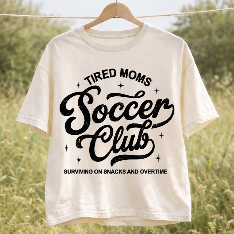 May include: Cream-colored t-shirt with the text "TIRED MOMS Soccer Club" in black, surrounded by stars. The phrase "SURVIVING ON SNACKS AND OVERTIME" is printed below the club name. The shirt is hanging on a wooden hanger.