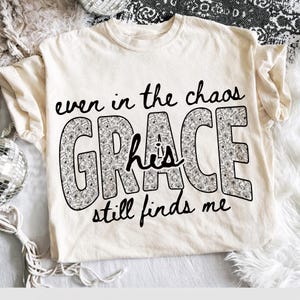 May include: Cream-colored t-shirt with the phrase "even in the chaos GRACE his still finds me" in black script. The word "GRACE" is filled with a floral pattern. The shirt has short sleeves and a crew neck.