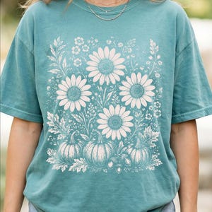 May include: A teal short-sleeve t-shirt featuring a large, cream-colored graphic print on the front. The design displays a collection of sunflowers, various wildflowers, three pumpkins, and an acorn arranged in a botanical illustration style.