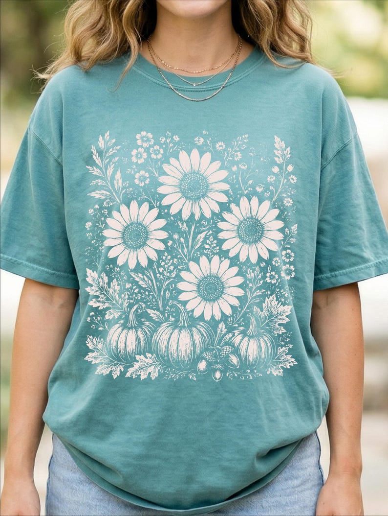 May include: A teal short-sleeve t-shirt featuring a large, cream-colored graphic print on the front. The design displays a collection of sunflowers, various wildflowers, three pumpkins, and an acorn arranged in a botanical illustration style.