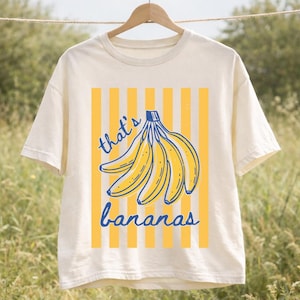May include: Cream-colored t-shirt with a yellow and blue banana graphic. The design features a bunch of bananas with the text "that's bananas" in blue script. The shirt is hanging on a wooden hanger.