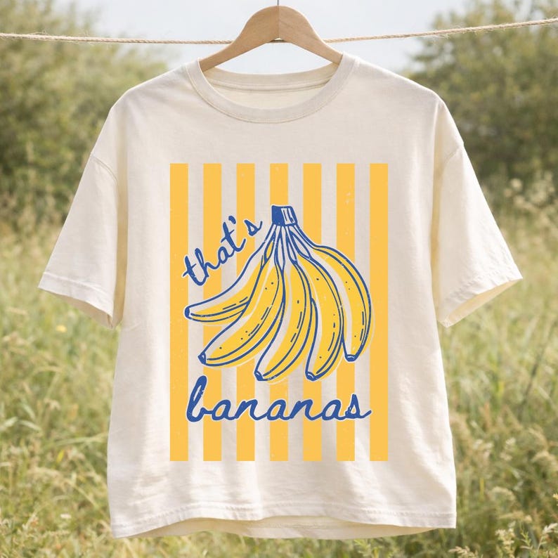 May include: Cream-colored t-shirt with a yellow and blue banana graphic. The design features a bunch of bananas with the text "that's bananas" in blue script. The shirt is hanging on a wooden hanger.