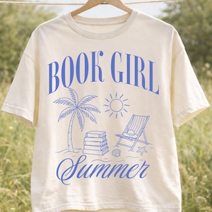 Op de afbeelding: Crèmekleurig T-shirt met de woorden "BOOK GIRL Summer" in blauw. Het ontwerp bevat een palmboom, zon, boeken, strandstoel en zeeschelp. Het shirt hangt aan een houten hanger.
