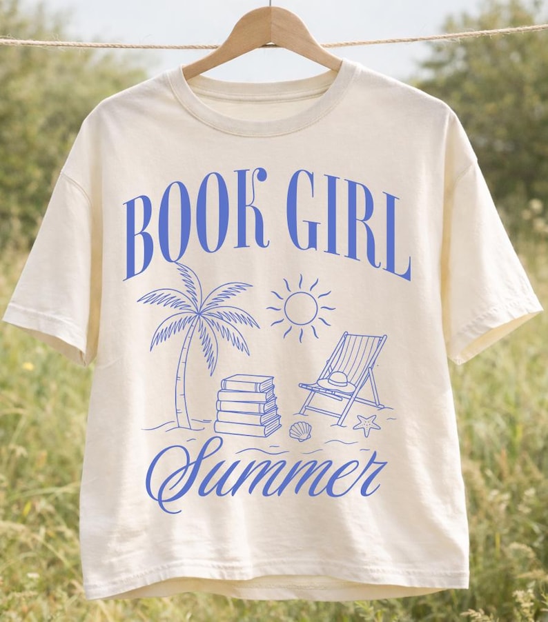 May include: Cream-colored t-shirt with the words "BOOK GIRL Summer" in blue script. The design includes a palm tree, sun, books, beach chair, and seashell. The shirt is hanging on a wooden hanger.