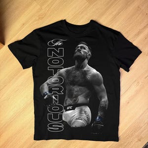 May include: Black t-shirt featuring a black and white image of a man with tattoos, wearing boxing gloves and shorts. The text "The NOTORIOUS" is printed vertically on the left side of the shirt. The UFC logo is visible on the shorts.