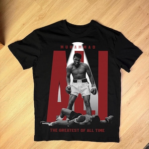 May include: Black t-shirt featuring a black and white image of Muhammad Ali standing over a defeated opponent. The word "ALI" is in large red letters, with the text "MUHAMMAD" above and "THE GREATEST OF ALL TIME" below.