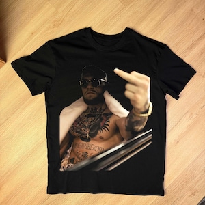 May include: Black t-shirt featuring a shirtless man with tattoos, sunglasses, and a gold bracelet. The man is giving the middle finger. The word "McGREGOR" is visible on the chest. The shirt is made of a soft material.