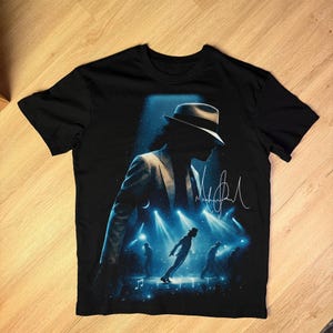 May include: Black t-shirt featuring a silhouette of a performer in a fedora and suit, with a signature and stage lights. The design includes figures dancing on a stage with musical notes.