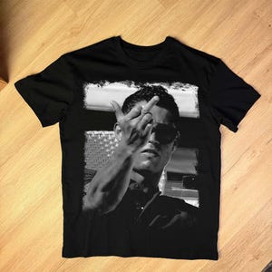 May include: Black t-shirt featuring a black and white image of a person wearing sunglasses, raising their middle finger. The image is framed with a white border. The t-shirt is made of a soft, comfortable material.
