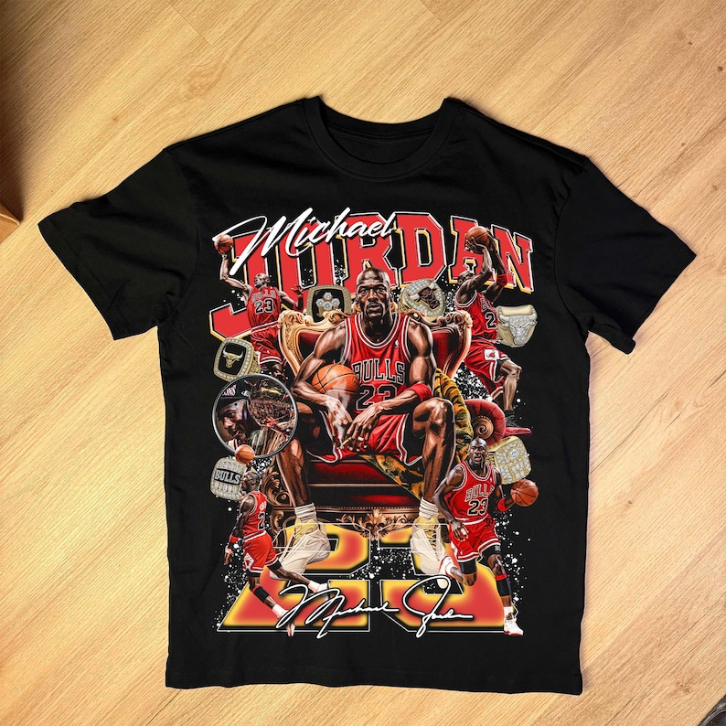May include: Black t-shirt featuring a graphic of Michael Jordan in a red Chicago Bulls uniform, seated on a throne. The design includes the text "Michael Jordan" and his signature, along with images of basketballs and championship rings.