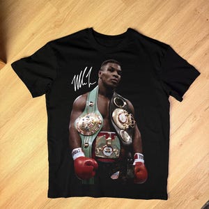 May include: Black t-shirt with a portrait of Mike Tyson, wearing boxing championship belts and red gloves. The shirt features Tyson's signature in white. The image is a vibrant depiction of the boxing legend.