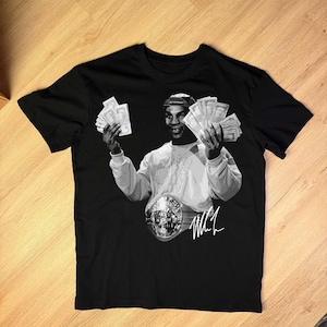May include: Black t-shirt with a black and white image of a boxer holding cash and wearing a championship belt. The image includes the boxer's signature. The shirt is made of a soft material.