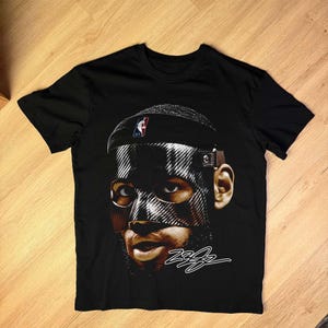 May include: Black t-shirt featuring a graphic of a person wearing a mask and a headband with an NBA logo. The image has a signature in white. The shirt is made of a soft material.