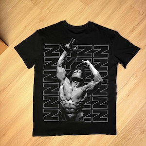 May include: Black t-shirt featuring a grayscale image of a muscular person with arms raised. The person is making a peace sign with one hand. The word "NYNN" is printed vertically in white on the left side of the image.