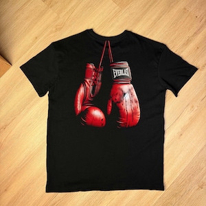 Puede incluir: Camiseta negra con un gráfico de dos guantes de boxeo rojos. Los guantes están detallados con cordones y la palabra "EVERLAST" en el puño. El diseño tiene una apariencia realista y ligeramente desgastada.