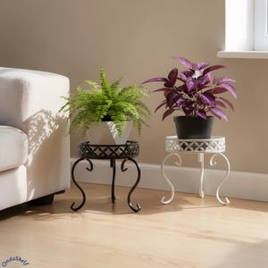 May include: Two decorative plant stands, one black and one white, each supporting a potted plant. The black stand holds a green fern in a white geometric pot. The white stand holds a purple plant in a black pot. Both stands have ornate, scrolled legs.