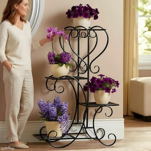 May include: A black wrought iron plant stand with multiple tiers, holding various potted plants with purple flowers. The stand has a decorative heart-shaped design and is placed indoors.
