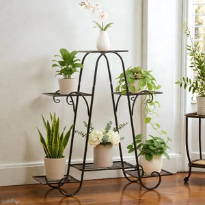 Może przedstawiać: Czarny, metalowy stojak na rośliny z wieloma poziomami, na którym znajdują się różne rośliny doniczkowe i biały wazon z kwiatami. Stojak ma dekoracyjny, kręcony design i jest umieszczony w pobliżu okna.