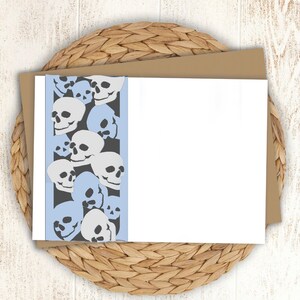 May include: A white card with a light blue border and a pattern of black and white skulls. The card is folded in half and the front is blank.