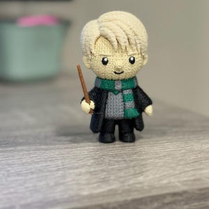 May include: A handmade, crocheted doll of a character with blonde hair, wearing a black robe, a green and gray scarf, and holding a brown wand. The doll has a smiling face and is standing on a gray surface.