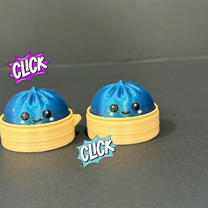 May include: Two blue, shiny, rounded dumpling figures with smiling faces, each resting inside a light tan, circular, tiered steamer basket. The word CLICK is displayed in purple and blue comic-style speech bubbles near the figures.