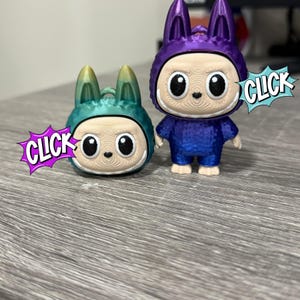 Puede incluir: Dos figuras impresas en 3D, una turquesa y amarilla, la otra morada y azul, ambas parecidas a un personaje con un disfraz de conejito. Las figuras tienen ojos grandes y una boca sonriente. La palabra "CLICK" está en una burbuja.