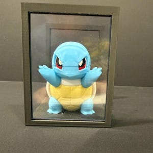May include: A framed 3D-printed figure of a Squirtle character. The figure is light blue with a yellow shell and red eyes, encased in a black frame. The artwork is displayed against a dark background.