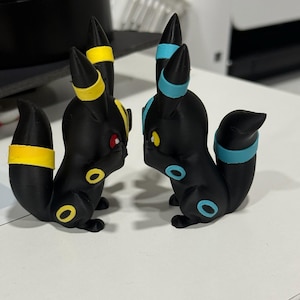 May include: Two black fox-like figurines facing each other. The left figurine features yellow rings on its ears, tail, and legs with red eyes. The right figurine features light blue rings on its ears, tail, and legs with yellow eyes. Both have pointed ears.