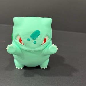 Puede incluir: Figura de Bulbasaur impresa en 3D, de color verde claro. Tiene ojos rojos, una mancha verde en la frente y garras blancas. La figura está sobre una superficie oscura.