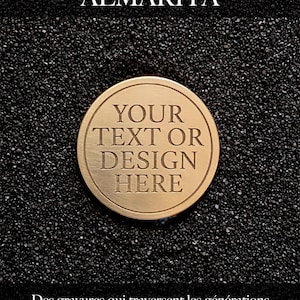 Op de afbeelding: Een ronde goudkleurige metalen munt met de gegraveerde tekst "YOUR TEXT OR DESIGN HERE" in het midden. Boven de munt staat de tekst "Atelier ALMARITA" en daaronder de Franse zin "Des gravures qui traversent les générations et célèbrent l'essentiel."