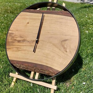 Könnte beinhalten: Eine kreisförmige Wanduhr mit einem hellbraunen Holzgehäuse, das eine natürliche Baumkante und eine dunkle Rindenstruktur an der Unterseite aufweist. Die Uhr ist von einem dünnen schwarzen Metallrahmen umgeben und hat zwei schmale schwarze Zeiger.