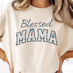May include: Cream-colored t-shirt with the words "Blessed MAMA" printed on the front. The word "MAMA" is in a collegiate-style font, filled with a floral pattern in shades of blue, pink, and white. The text is in a dark blue color.