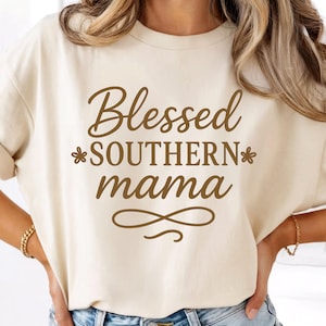 May include: Cream-colored t-shirt with the words "Blessed SOUTHERN mama" in brown script. The text is centered and includes floral accents and a decorative flourish. The shirt is a casual style.