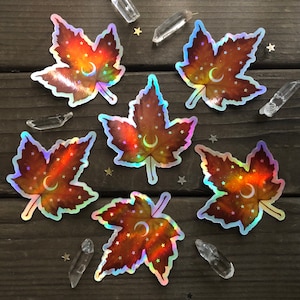May include: Six iridescent holographic stickers of maple leaves with a crescent moon and stars on a brown wooden background.