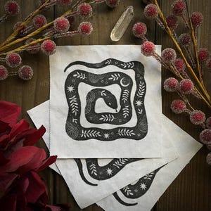 May include: A set of three white fabric coasters with a black and white illustration of a snake curled into a square shape. The snake is surrounded by small stars and leaves.
