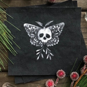 May include: A black fabric patch with a white design of a skull inside a butterfly with floral details.
