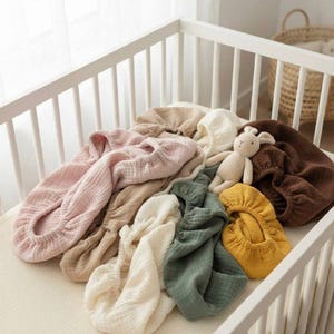 Puede incluir: Una colección de fundas de tela de muselina suave y texturizada en varios colores, incluyendo rosa, beige, crema, marrón oscuro, amarillo mostaza y verde salvia, acompañada de un pequeño conejo de punto de color claro con orejas largas.