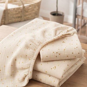 May include: A cream-colored fitted sheet made of soft, textured muslin fabric featuring a pattern of small, metallic gold polka dots. The sheet is folded into a neat stack, showing the elasticized edge and the lightweight, breathable material.
