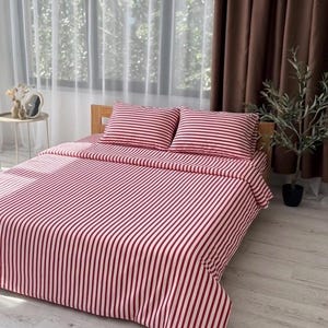Op de afbeelding: Een bed met een rood-witte gestreepte beddengoedset. De set bevat een hoeslaken, een laken en twee kussenslopen. Het bed is van hout en staat in een lichte kamer met een raam en gordijnen.